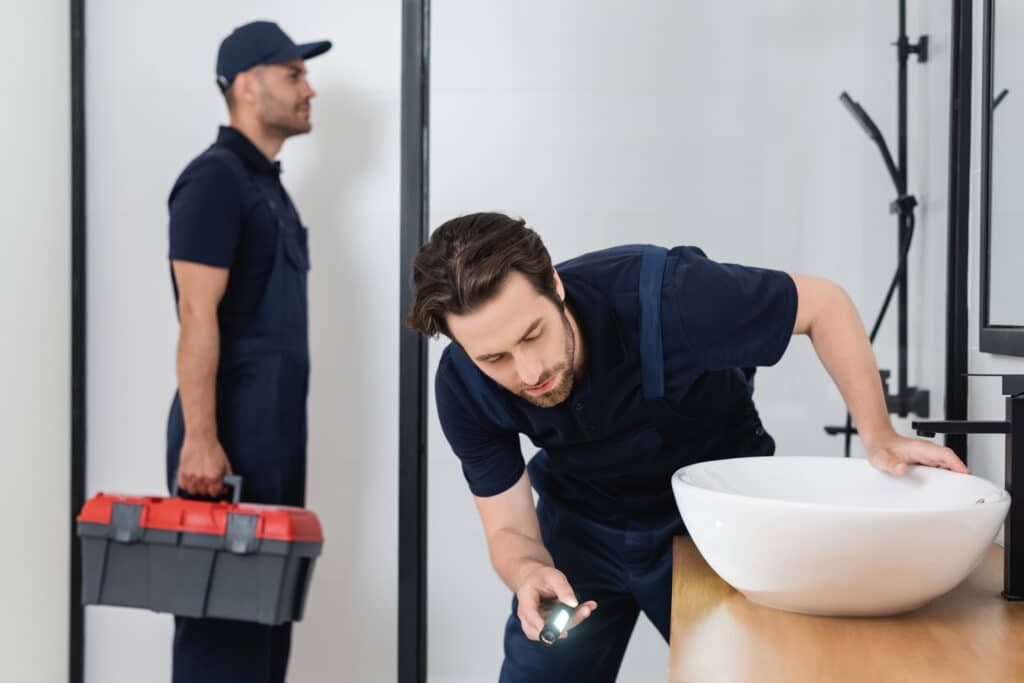 plumber with flashlight checking sink in bathroom near blurred colleague with toolbox